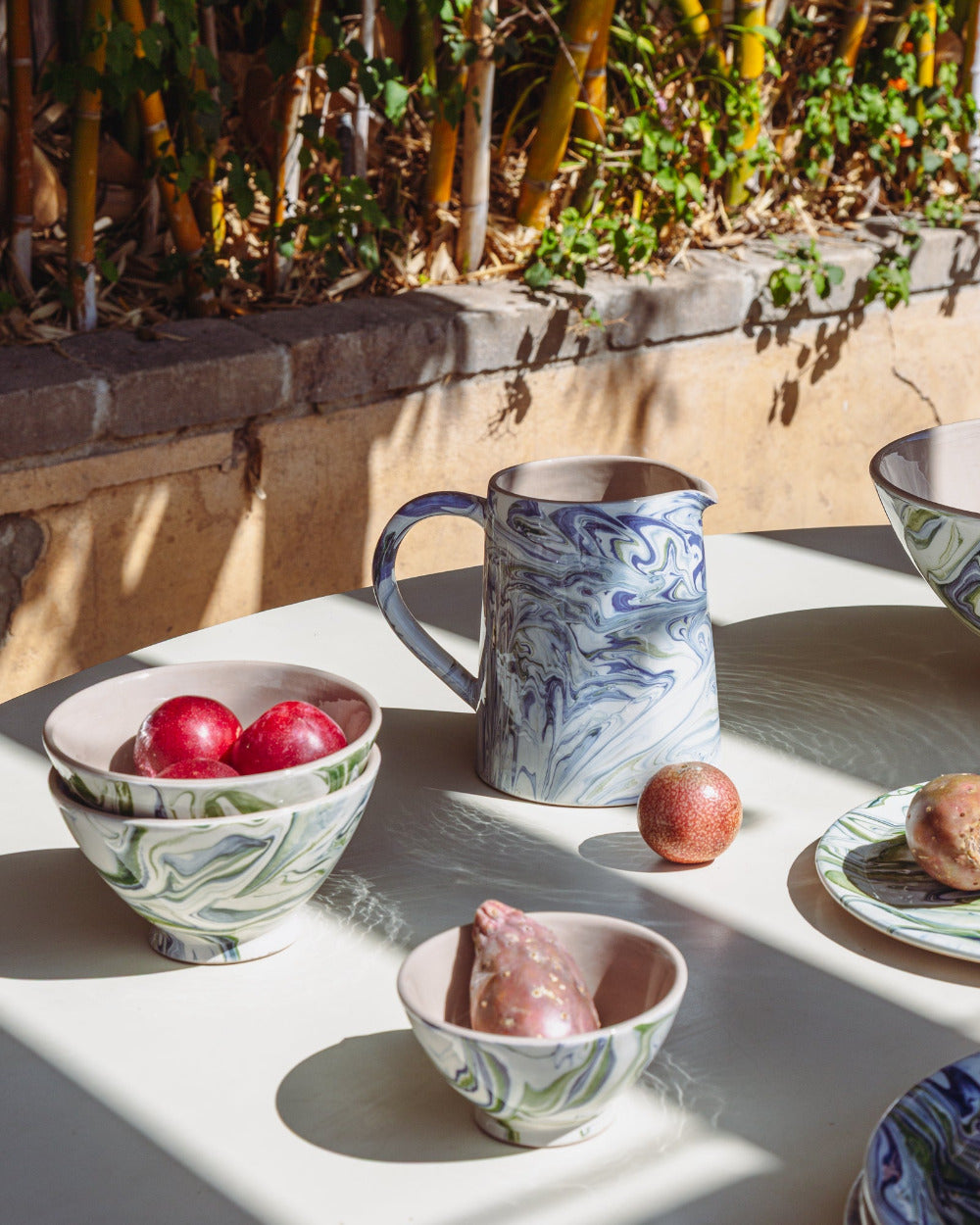 Ceramic bowls and a pitcher on a table outdoors with plants in the background