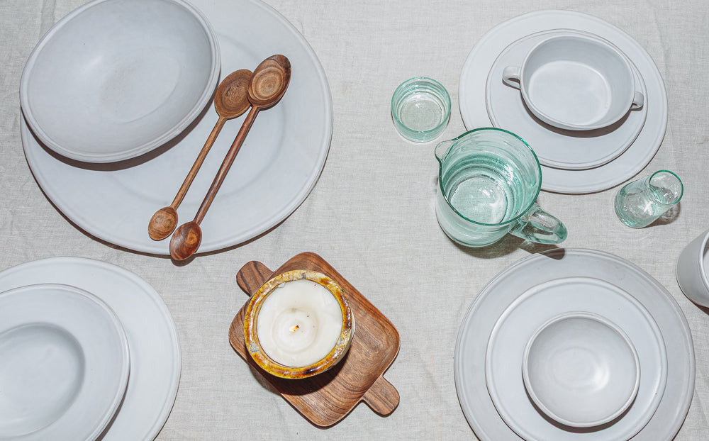 Dining table setting with white plates, green glasses, wooden spoons, and a candle on a wooden coaster.