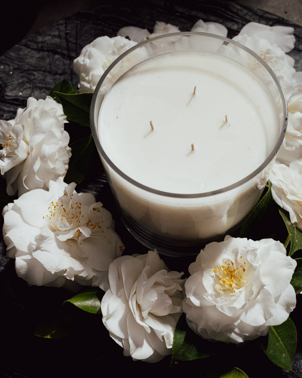 White candle in a glass jar surrounded by white flowers on a dark surface