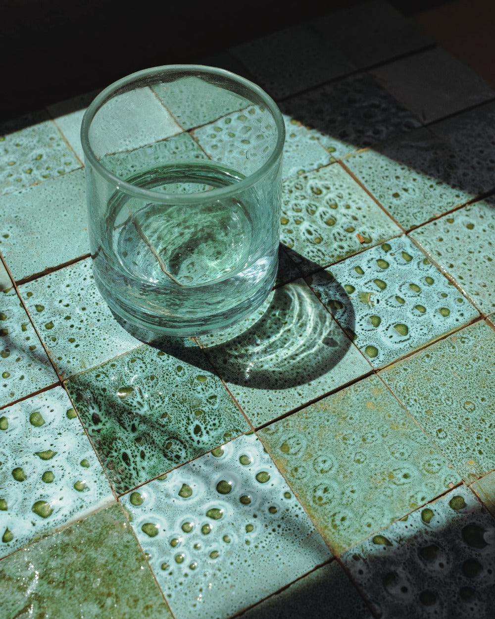 Clear glass of water on a textured green, reactive glaze zellige tile surface