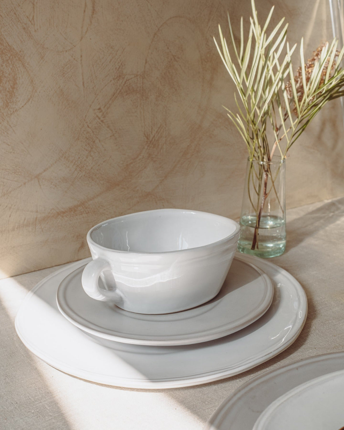 White ceramic bowl and plates on a neutral background with a vase of plants.