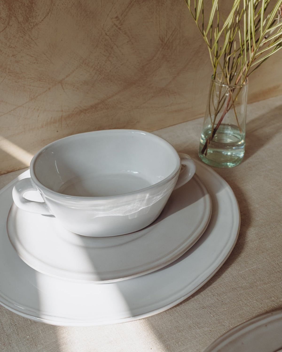 White ceramic bowl and saucer on a wooden surface with a glass of water and plant in the background.