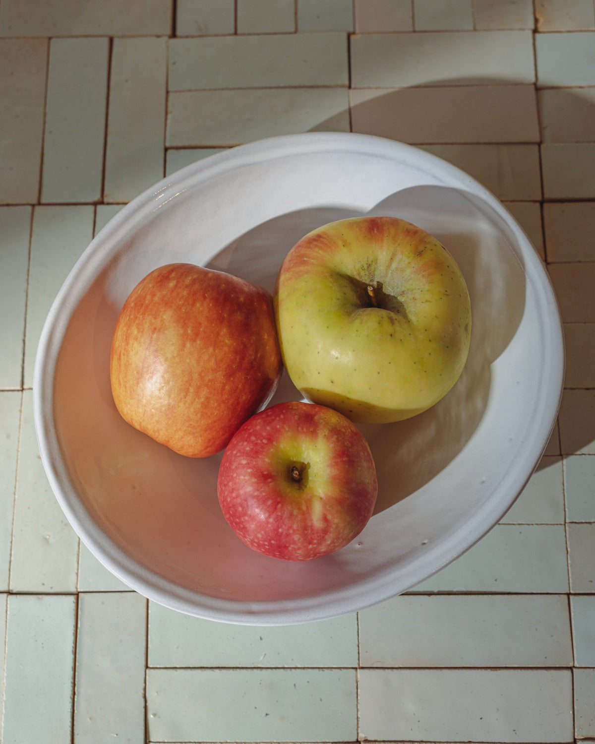 Three apples in a white oval bowl on a tiled table