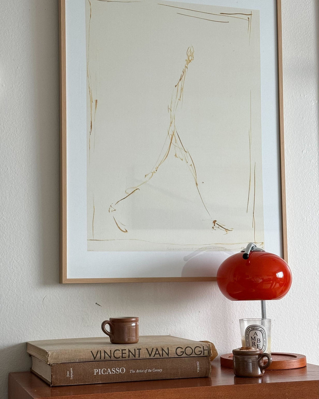 Framed artwork on a wall above a wooden cabinet with french espresso cups, books and decorative items.