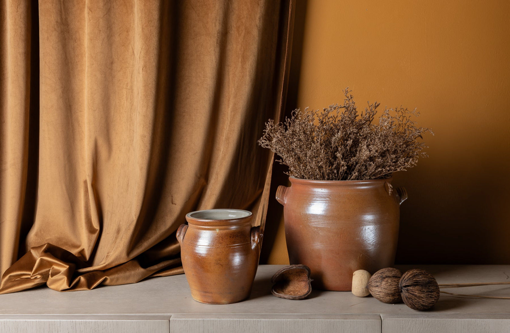 Two vintage, french salt glazed ceramic pots with dried plants and nuts on a wooden surface against a caramel colored curtain.