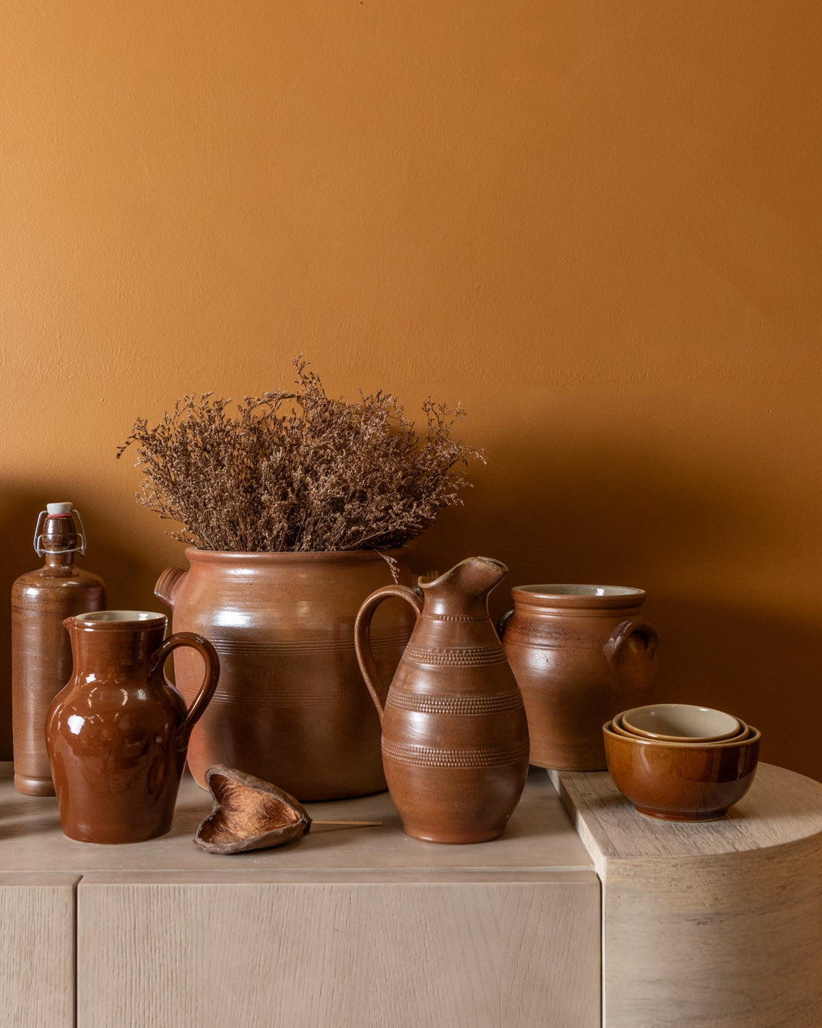 Collection of brown ceramic pots and vases on a wooden surface against an orange wall.